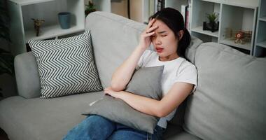 Handheld high angle view shot, Asian woman sitting alone on the sofa, looking sad, hold the head with hand while thinking about something in the living room at home. Facial expression concept. video
