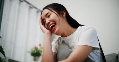 Handheld low angle view shot, Young woman is sitting on a sofa, smiling happily while thinking about something in the living room at home. video