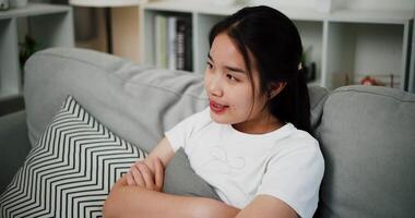 Handheld high angle view shot, Attractive young woman is sitting on a sofa, smiling happily while thinking about something in the living room at home. video