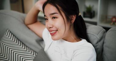 Handheld low angle view shot, Attractive young woman is sitting on a sofa, smiling happily while thinking about something in the living room at home. video