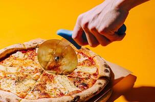 Hand using pizza cutter to slice fresh pizza on orange background photo