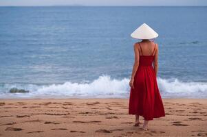 Vietnamese lady in traditional conical Non La hat and red dress on the beach against the background of the sea photo