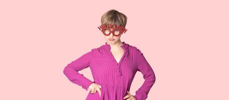 A cheerful young woman in Christmas glasses poses on a workspace layout highlighted on a simple pastel light pink background. photo
