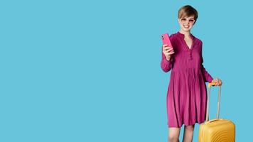 A young woman with a suitcase and a mobile phone in her hands, on a simple blue background. Copying the space. photo