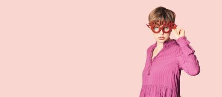 A cheerful young woman in Christmas glasses poses on a workspace layout highlighted on a simple pastel light pink background. photo