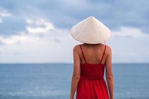 Vietnamese woman in traditional conical Non La hat and red dress on the background of the sea. View from the back. photo
