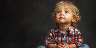 Young child with curly hair gazes upward while seated in a plaid shirt against a dark backdrop photo