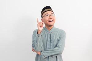 Asian Muslim man smiling doing pointing up gesture in Ramadan Eid Al Fitr and eid adha days celebration isolated white background. Islamic celebration concept. photo