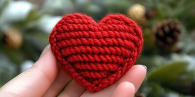 Handmade red knitted heart held in hand against a backdrop of greenery and pine cones during winter season photo