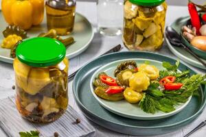 Pickled pattypan squash, gherkins, chili pepper and herbs served on a plate, with jars of pickles and other ingredients in the background photo