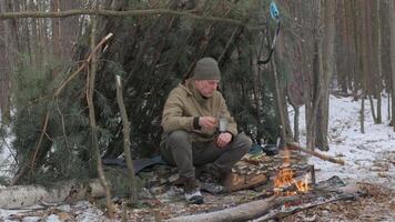 A man sits by a campfire in a winter forest. He has built a shelter from pine branches and is dressed in warm outdoor clothing. The scene reflects survival, bushcraft skills. video