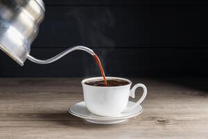 Pouring hot coffee into a white coffee cup With roasted coffee beans on a wooden table, dark black background. photo
