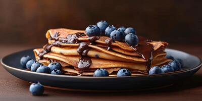 Delicious stack of pancakes topped with fresh blueberries and rich chocolate syrup served on a dark plate photo