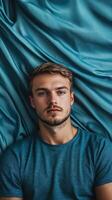 Young man in a blue t-shirt lying on textured blue fabric looking contemplative indoors during the day photo
