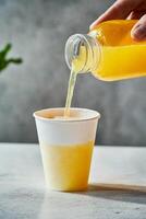 Refreshing lemonade is poured from a bottle into a white paper cup on a light gray concrete table photo