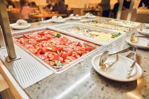 Buffet bar or buffet in the cafe. A metal container with sliced fresh tomatoes. Against the background of unrecognizable people dining photo
