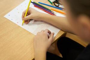 An unrecognizable child is intently drawing on a white sheet at a table with a set of colored pencils. With space to copy photo