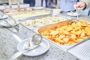 Close-up of a metal container with baked hearty food. A woman puts food in a cup on a buffet table in a restaurant. With space to copy photo