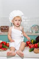 A little boy in a chef's outfit sitting on a counter with a bunch of vegetables photo