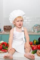 A little boy in a chef's hat sitting on a counter with a bunch of vegetables photo