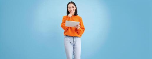 Woman with digital tablet thinking, looking aside with thoughtful face expression, standing over blue background photo