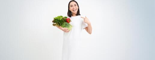 Image of happy korean woman, pointing at vegetables, smiling, recommending eating healthy raw food, green diet concept, white background photo