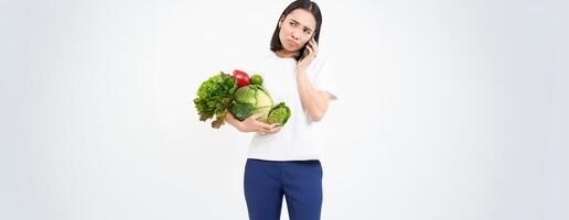 Image of girl with vegetables, making phone call with sad, unhappy face, isolated on white background photo