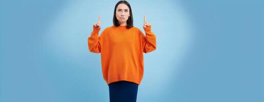 Perplexed asian girl, looks and points up with frustrated, thinking face expression, shows advertisement and reads it with doubt, stands over blue background photo
