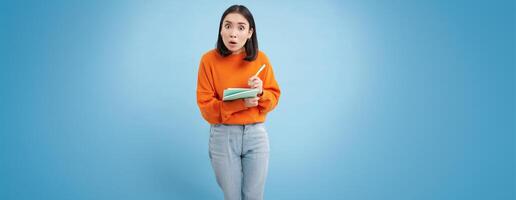 Education and students. Happy asian woman, holding notebooks and laughing, smiling at camera, enjoys going to University or College, blue background photo