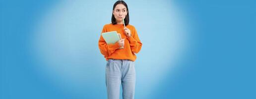 Asian girl, student with thinking face, pondering smth with serious face, holding pencil and notebooks, standing over blue background photo