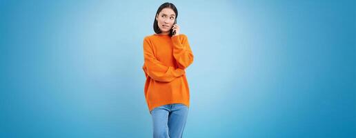 Girl with puzzled face answers phone call, looks confused while talks on cellphone, stands over blue background photo