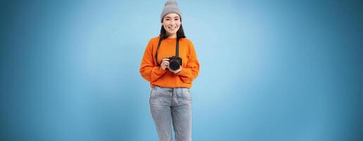Stylish asian girl with digital camera, taking pictures. Woman photographer smiling, standing over blue background photo