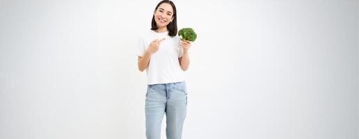 Portrait of cute asian woman, pointing at broccoli, showing vegetable, nutritionist recommending healthy diet food, white background photo