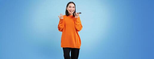 Cheerful smiling korean woman talk on speakerphone, records voice message, learns how to speak using language learning mobile app, blue background photo