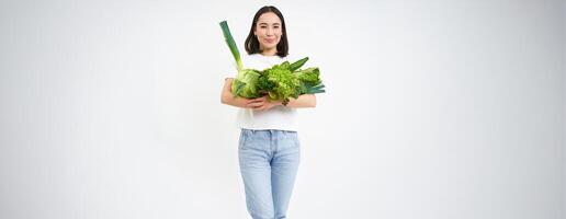 Portrait of happy young woman on green diet, likes eating organic food, holding fresh vegetables from garden, lettuce, cabbage, white studio background photo
