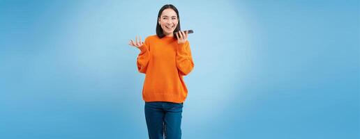 Happy asian woman records voice message, repeats words in language learning mobile app, talks in smartphone speakerphone, blue background photo