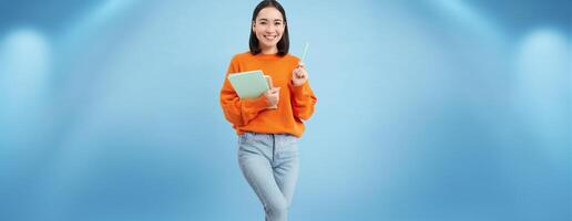 Education and students. Happy asian woman, holding notebooks and laughing, smiling at camera, enjoys going to University or College, blue background photo