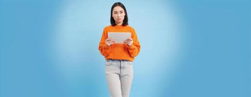 Woman with digital tablet thinking, looking aside with thoughtful face expression, standing over blue background photo