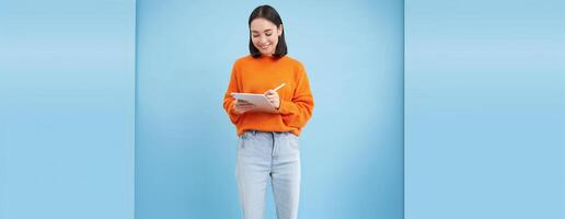 Portrait of smiling female tutor, girl student making notes on digital tablet, drawing in app, standing over blue background photo