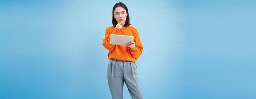 Woman with digital tablet thinking, looking aside with thoughtful face expression, standing over blue background photo