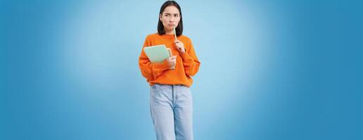 Asian girl, student with thinking face, pondering smth with serious face, holding pencil and notebooks, standing over blue background photo