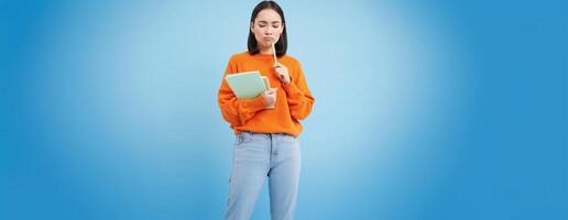Education and students. Happy asian woman, holding notebooks and laughing, smiling at camera, enjoys going to University or College, blue background photo