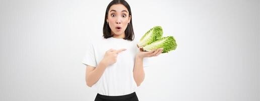 Portrait of surprised and amazed asian woman, points at cabbage, find out amazing benefit of lettuce, healthy vegetables, white background photo
