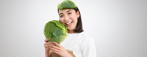 Close up portrait of healthy smiling asian woman, showing cabbage, green letture, with leaf on head, isolated on white background photo