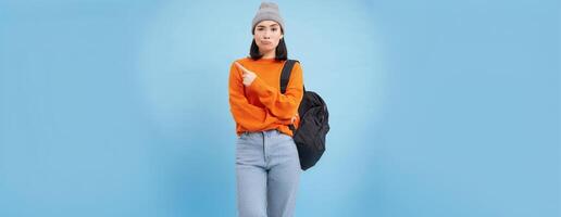 Skeptical asian woman pointing left at strange banner, looks doubtful, stands over studio background photo