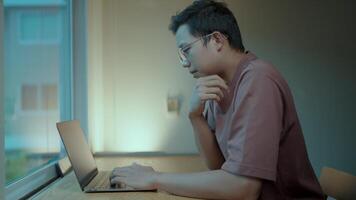 A young man sits at a table, thoughtfully working on his laptop. The soft light from a nearby window illuminates him in profile view.Displays a web page with a text interface using a modern chat tool. video