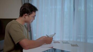 Man counting money at home. A young man sits at a table, meticulously counting money, his expression thoughtful and serious. The soft light from a nearby window illuminates the scene. video