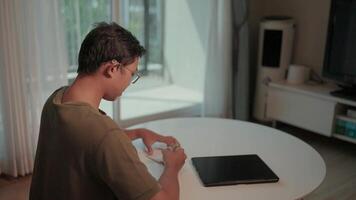 Focused on the task a man sits at a table, engrossed in working on his tablet and papers. The warm lighting of his home creates a calm and productive atmosphere. video