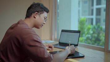 A young talented artist is captured in the act of creation, drawing on a digital tablet with a focused gaze. He sits by a window, sunlight filtering through the leaves. video