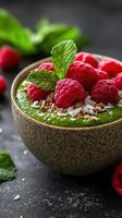 Delicious green smoothie bowl topped with fresh raspberries and mint leaves in a rustic bowl photo